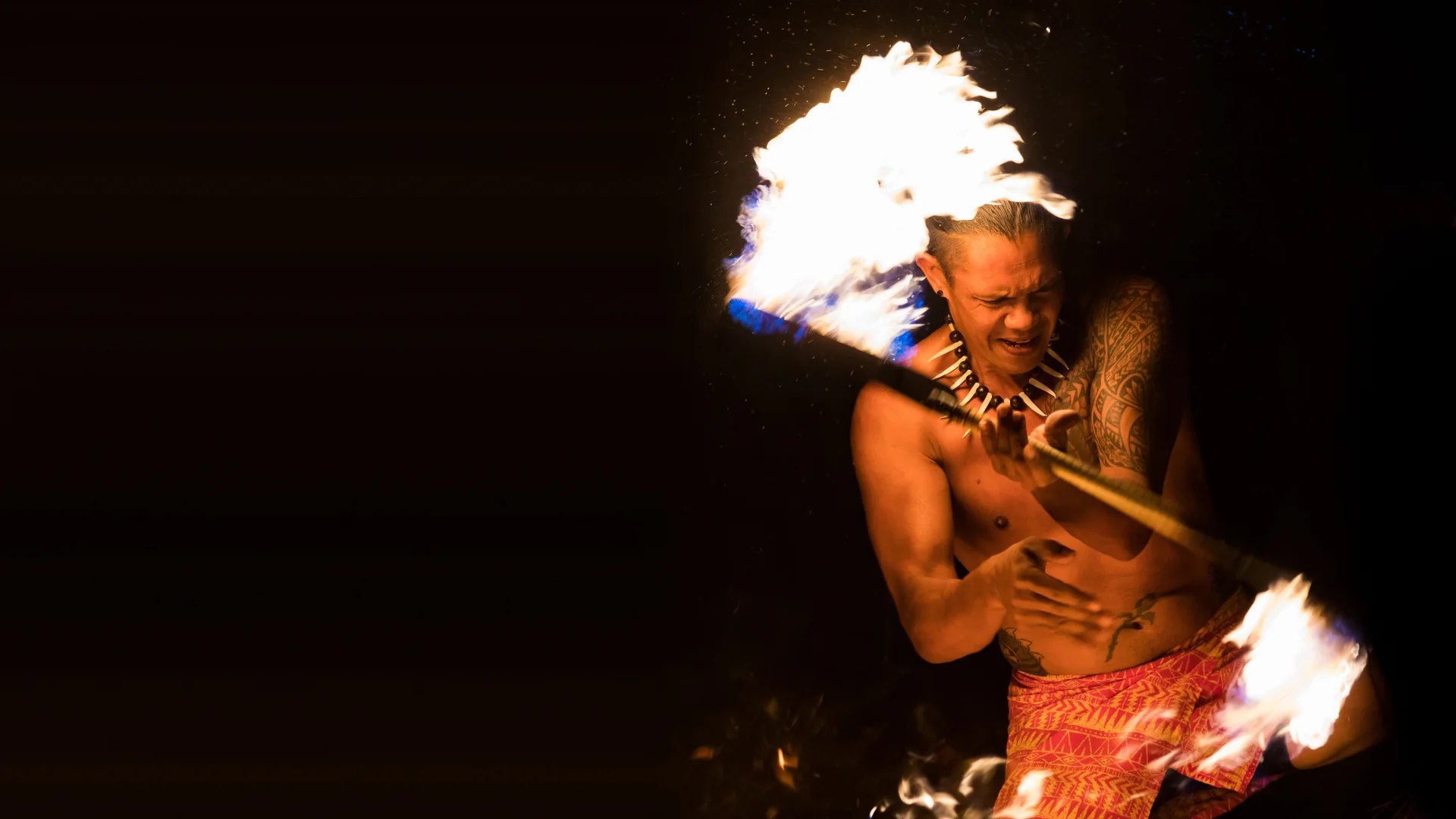 Um dançarino de faca em chamas em plena rotação no Luau Kalamaku, um bastão incandescente deixando um rasto de fogo contra a noite, usando um colar de dentes e calções vermelhos estampados