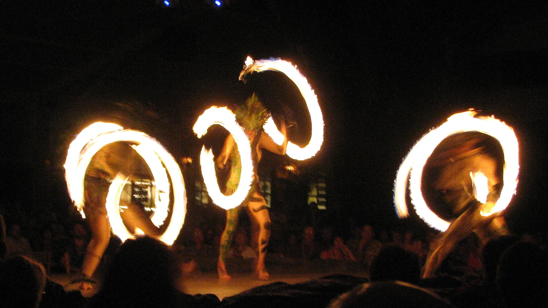 Três dançarinos de fogo no palco do Luau Kalamaku, cada um a girar um anel de chamas na escuridão enquanto o público assiste